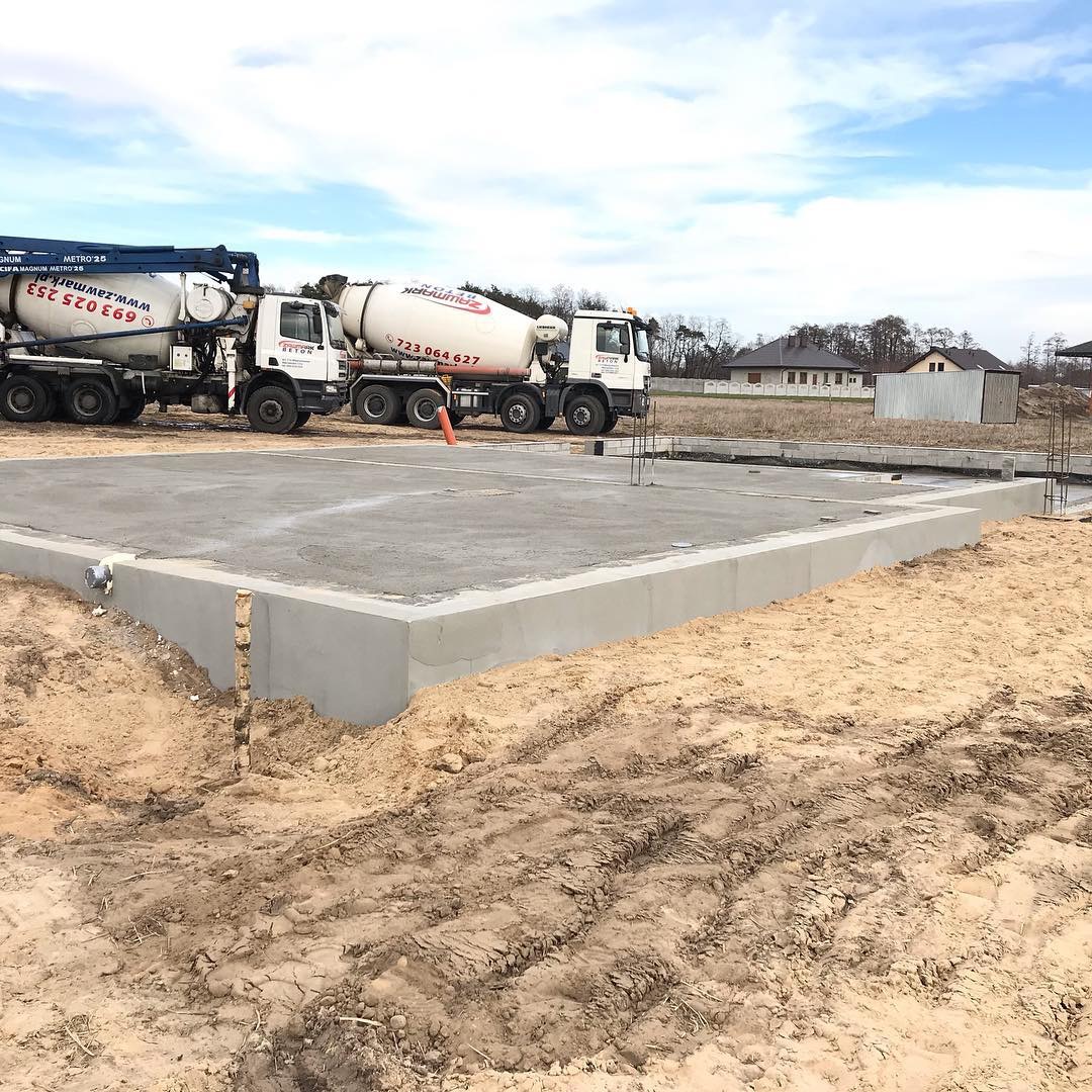 Wylana betonowa płyta fundamentowa z wystającymi prętami zbrojeniowymi, dwa betonomieszarki zaparkowane w tle, teren budowy z piaskiem i śladami opon.