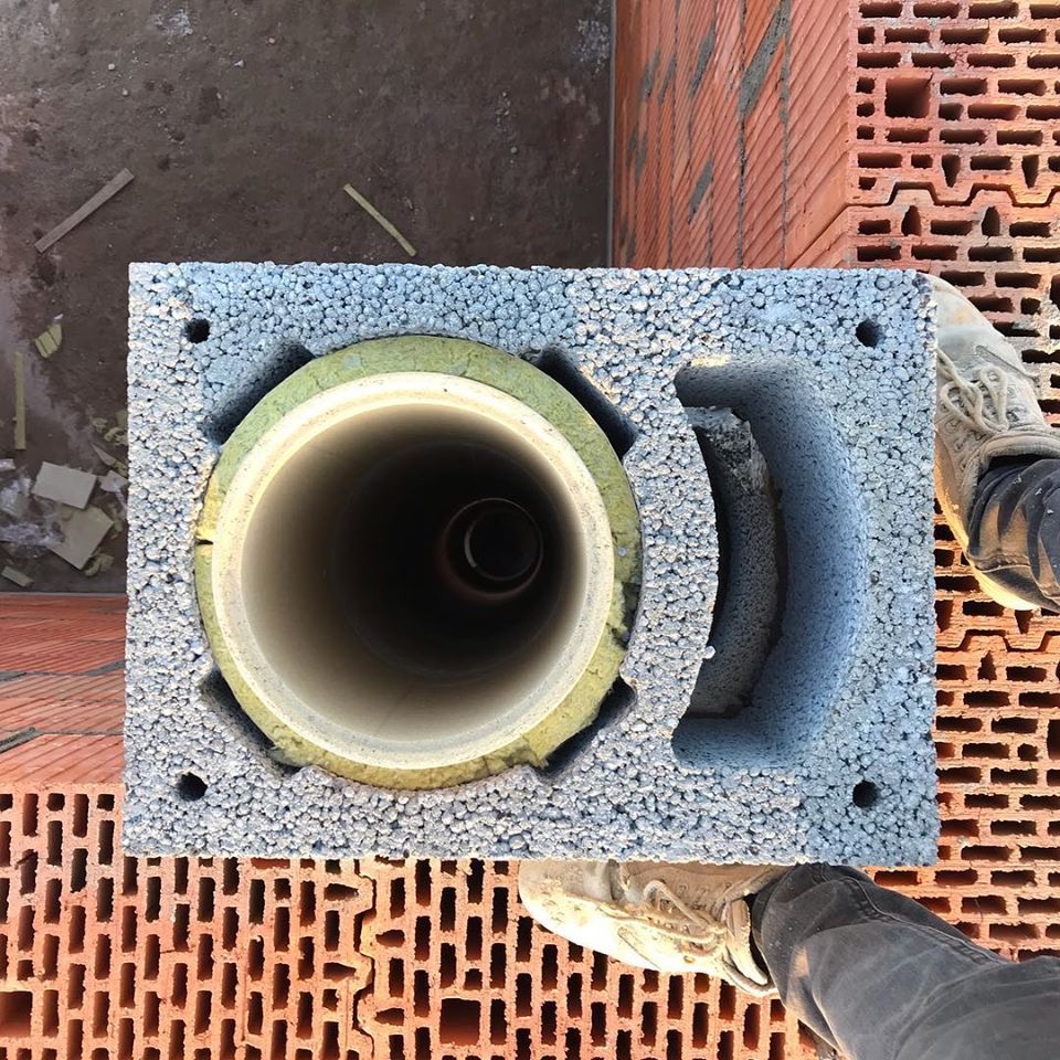 Wkład kominowy z izolacją umieszczony w bloczku z szarego styropianu, widok z góry na tle muru z czerwonej cegły dziurawki i fragmentów obuwia roboczego.