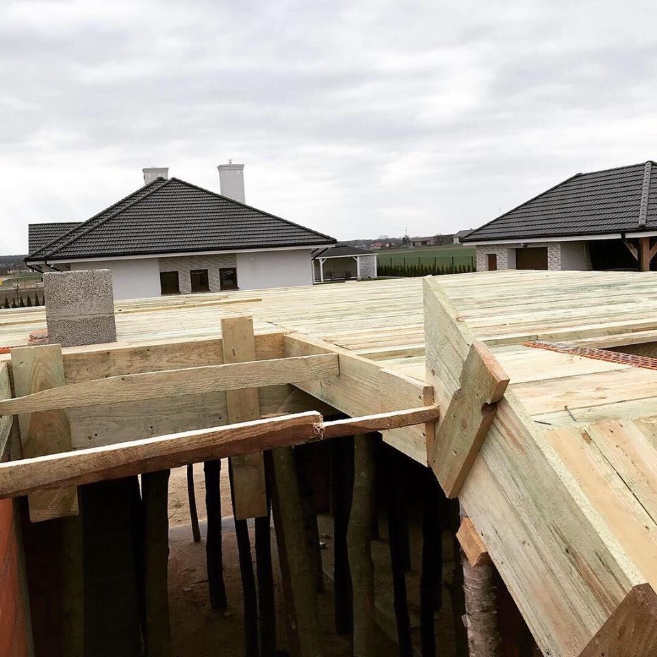 Budowa domu w trakcie wznoszenia konstrukcji drewnianej dachu, widoczne elementy szalunku i podparcia stropu, w tle domy z ciemną dachówką i kominami.