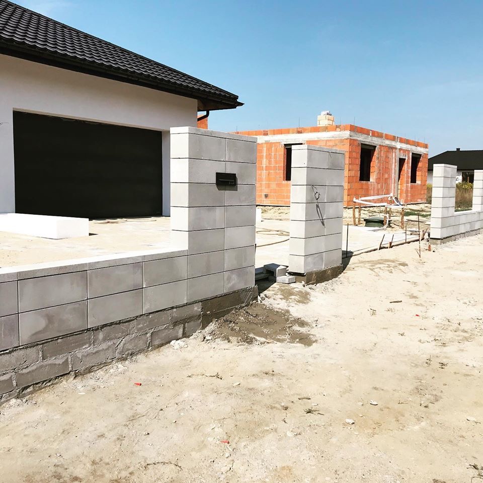 Budowa domu w trakcie realizacji: widoczny garaż z czarną bramą, fragment muru z szarej kostki z zamontowaną skrzynką na listy oraz budynek w stanie surowym otwartym z czerwonej cegły w tle.