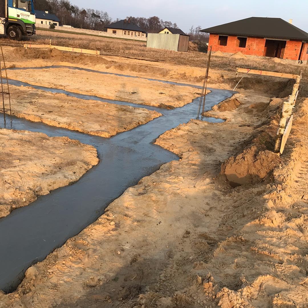 Wylane fundamenty pod budowę domu jednorodzinnego, widoczne zbrojenia i szalunki, w tle ceglany dom w stanie surowym otwartym i krajobraz wiejski w ciepłym świetle zachodzącego słońca.