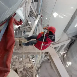 Pracownik w kasku i czerwonej kurtce, zabezpieczony linami, wykonuje prace na wysokości na konstrukcji przemysłowej, widok z dołu.