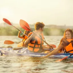 Trzy osoby w kajakach na wodzie, w pomarańczowych kamizelkach ratunkowych, wiosłują podczas słonecznego dnia.
