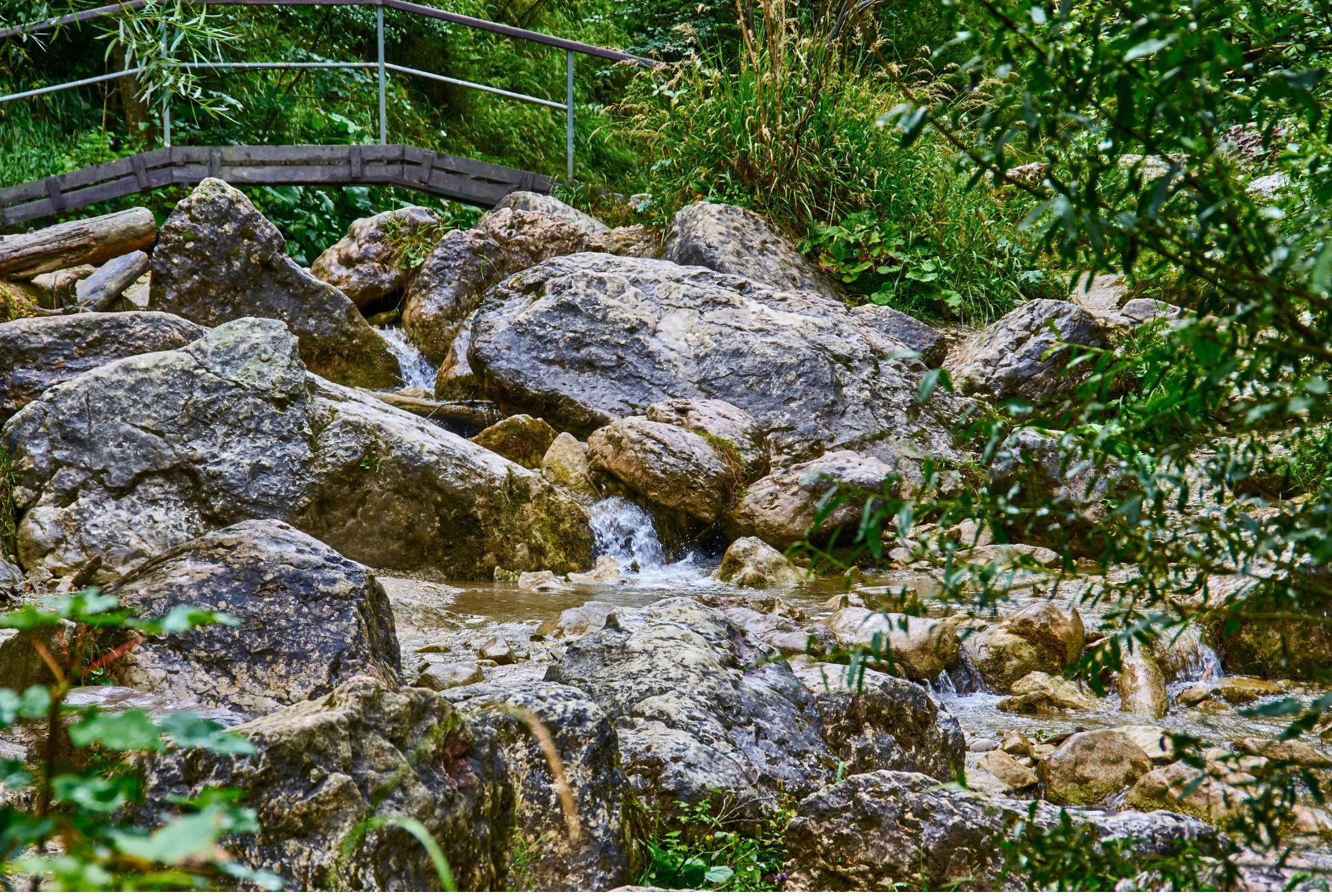 Kamienisty potok z kaskadami wody przepływający pod drewnianym mostkiem w otoczeniu zieleni.