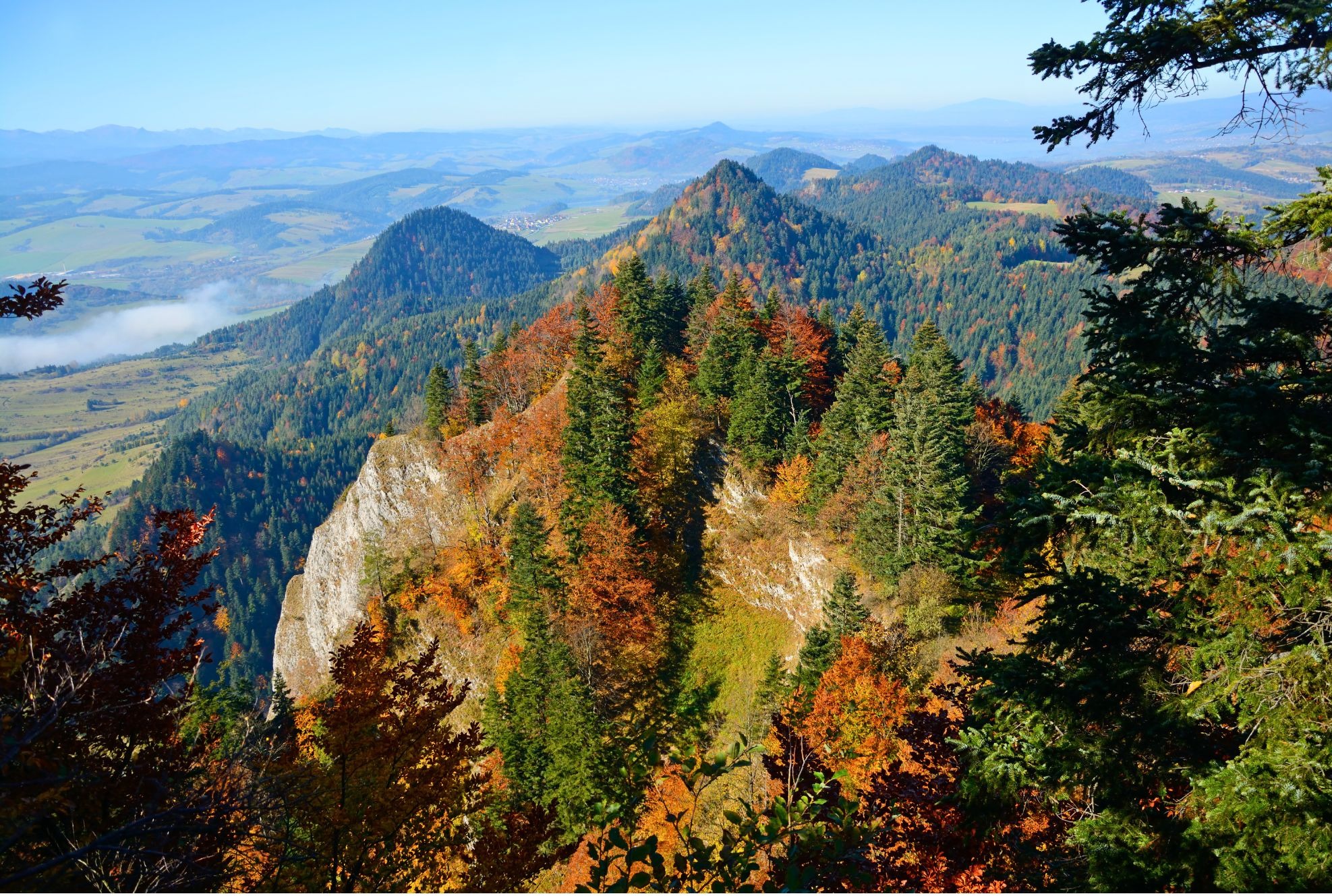 Jesienny krajobraz górski z widokiem na skalisty szczyt porośnięty drzewami iglastymi i liściastymi w barwach jesieni, w oddali dolina z mgłą i kolejne pasma gór.