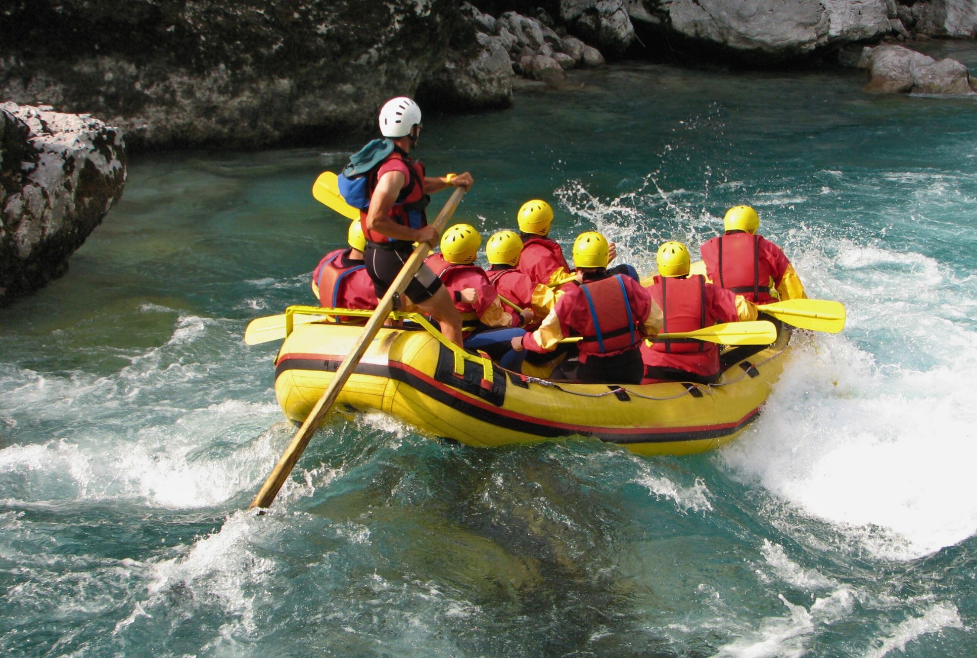Grupa w kaskach i kamizelkach płynie pontonem przez wzburzoną, turkusową rzekę z widocznymi skałami w tle.