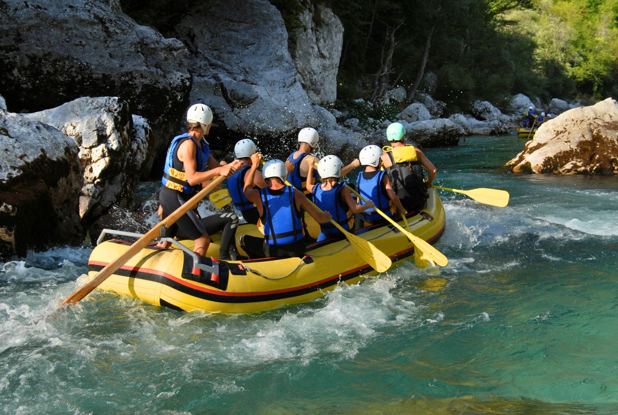 Żółty ponton z grupą osób w kaskach i kamizelkach płynie po rwącej, turkusowej rzece wśród skalistych brzegów i zieleni drzew.