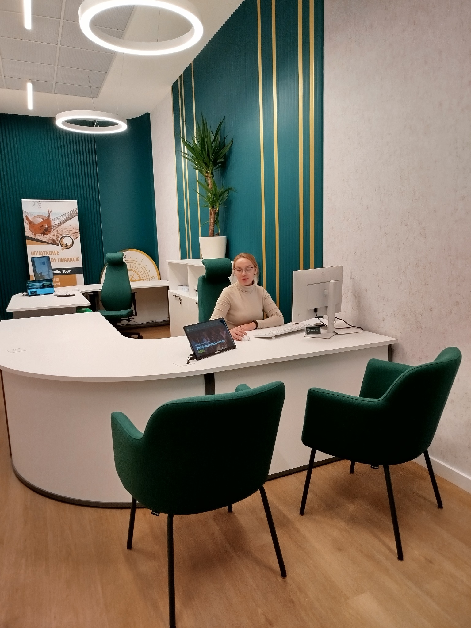 Wnętrze biura z recepcją w odcieniach zieleni i bieli, z pracownicą siedzącą za biurkiem i dwoma krzesłami dla klientów na pierwszym planie.