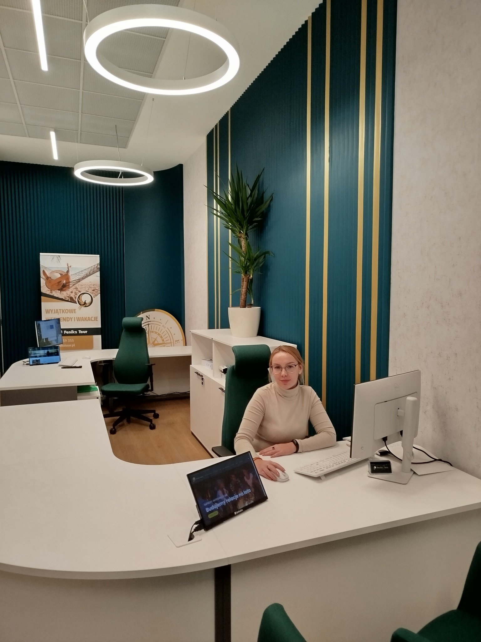 Wnętrze biura z pracownikiem siedzącym przy biurku, widoczny plakat z ofertą wyjazdów i wakacji oraz dekoracje ścienne w kolorze butelkowej zieleni z złotymi paskami.