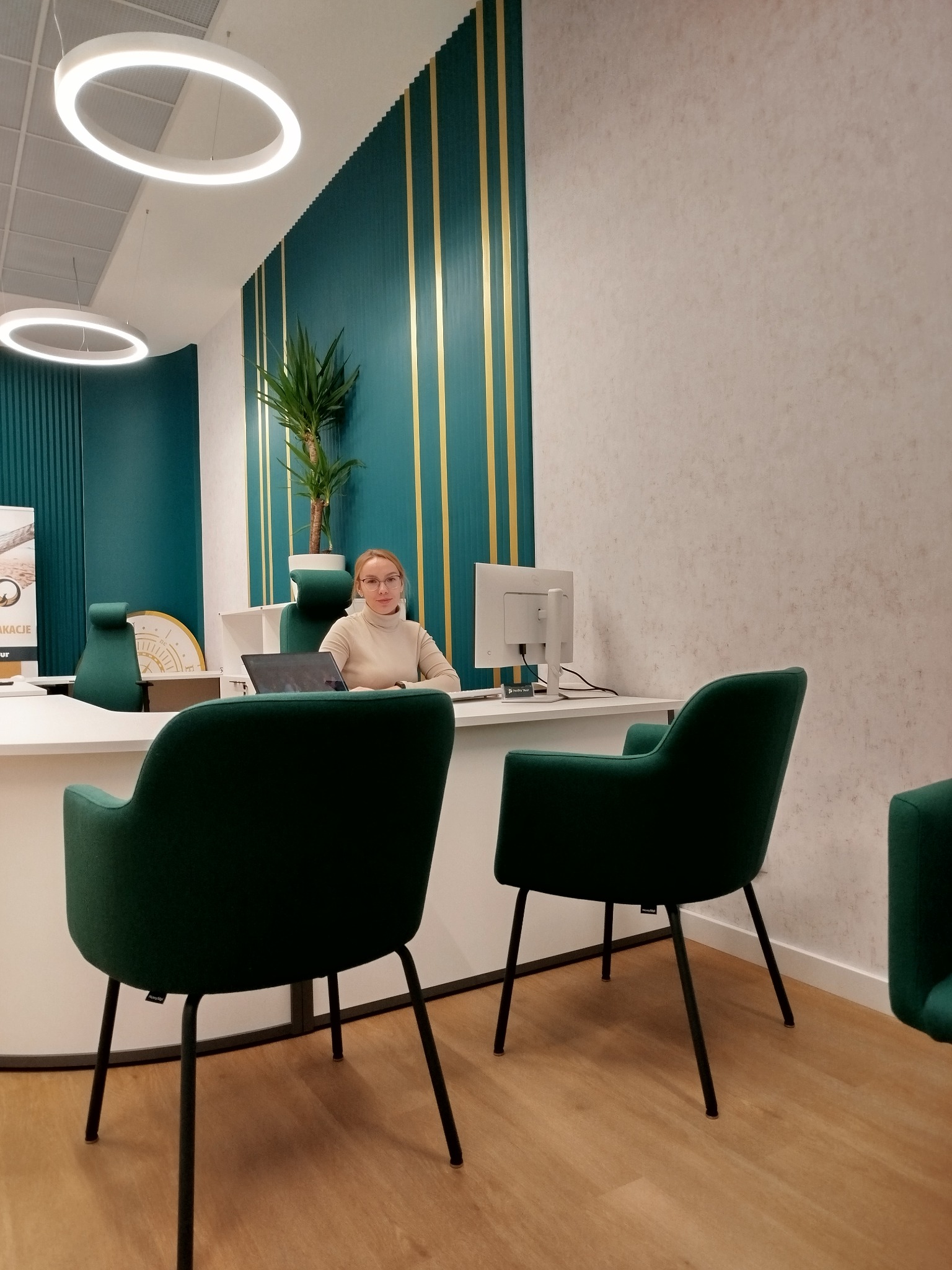 Pracownica biura podróży siedzi za biurkiem w nowoczesnym wnętrzu z zielonymi akcentami i okrągłymi lampami, czekając na klientów.