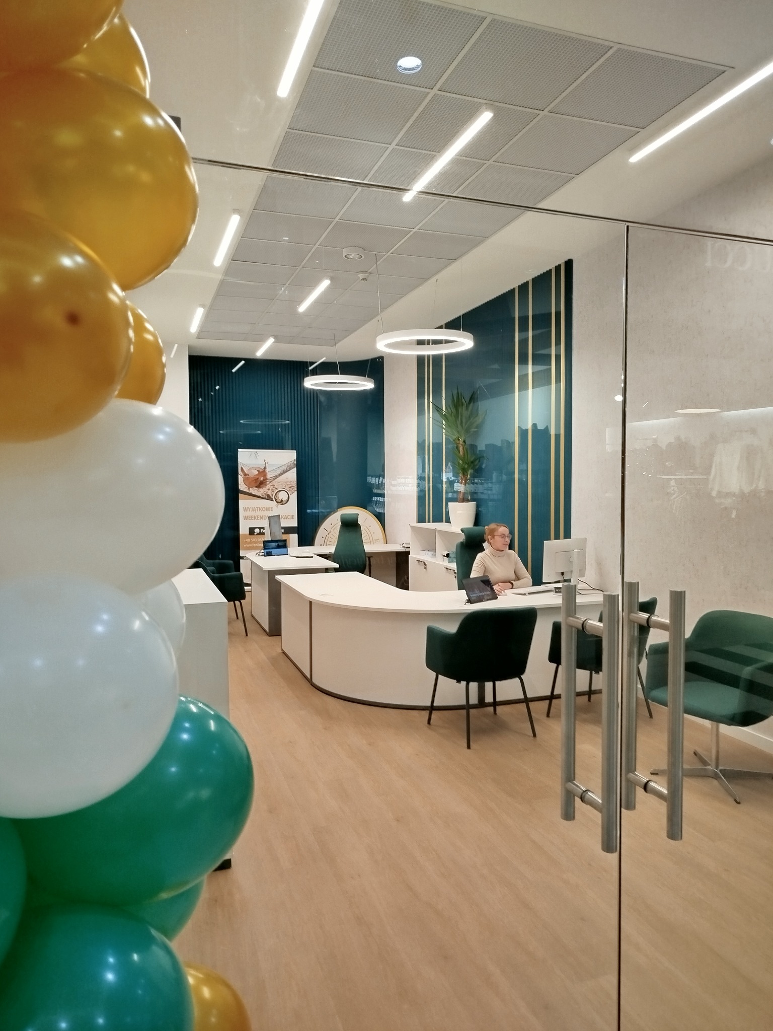 Wnętrze biura z recepcją, dekorowane balonami w kolorach złota, bieli i zieleni; pracownica siedzi za biurkiem, widoczny plakat z ofertą weekendowych wakacji.