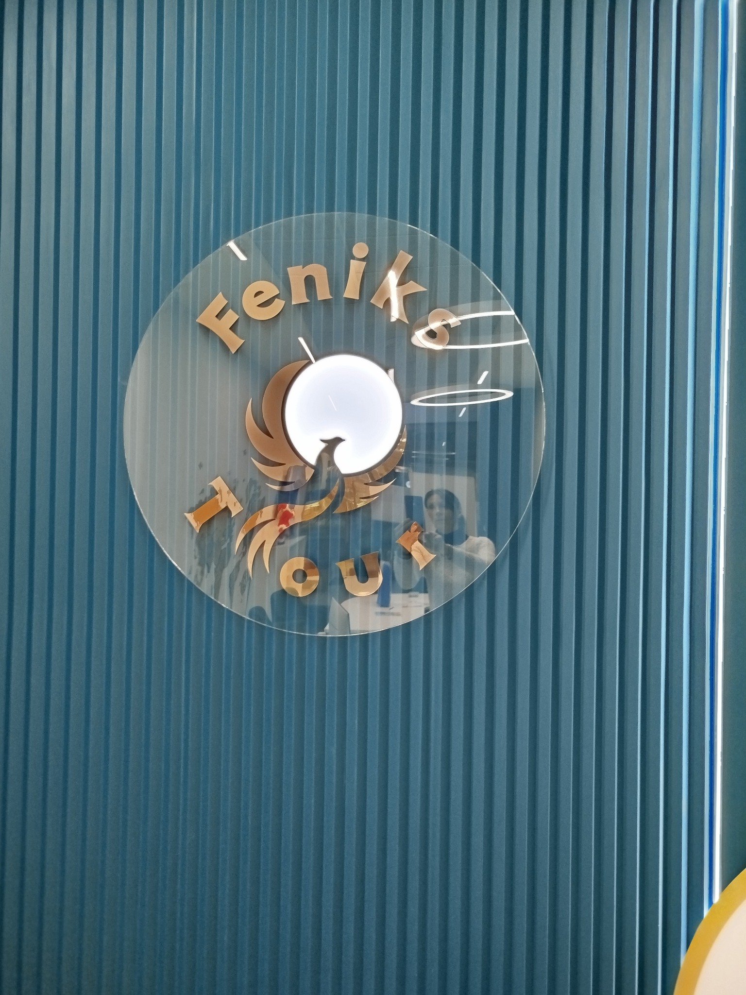 Okrągłe logo z napisem 'Feniks Tour' i stylizowanym wizerunkiem feniksa na tle niebieskiej ściany z pionowymi panelami.