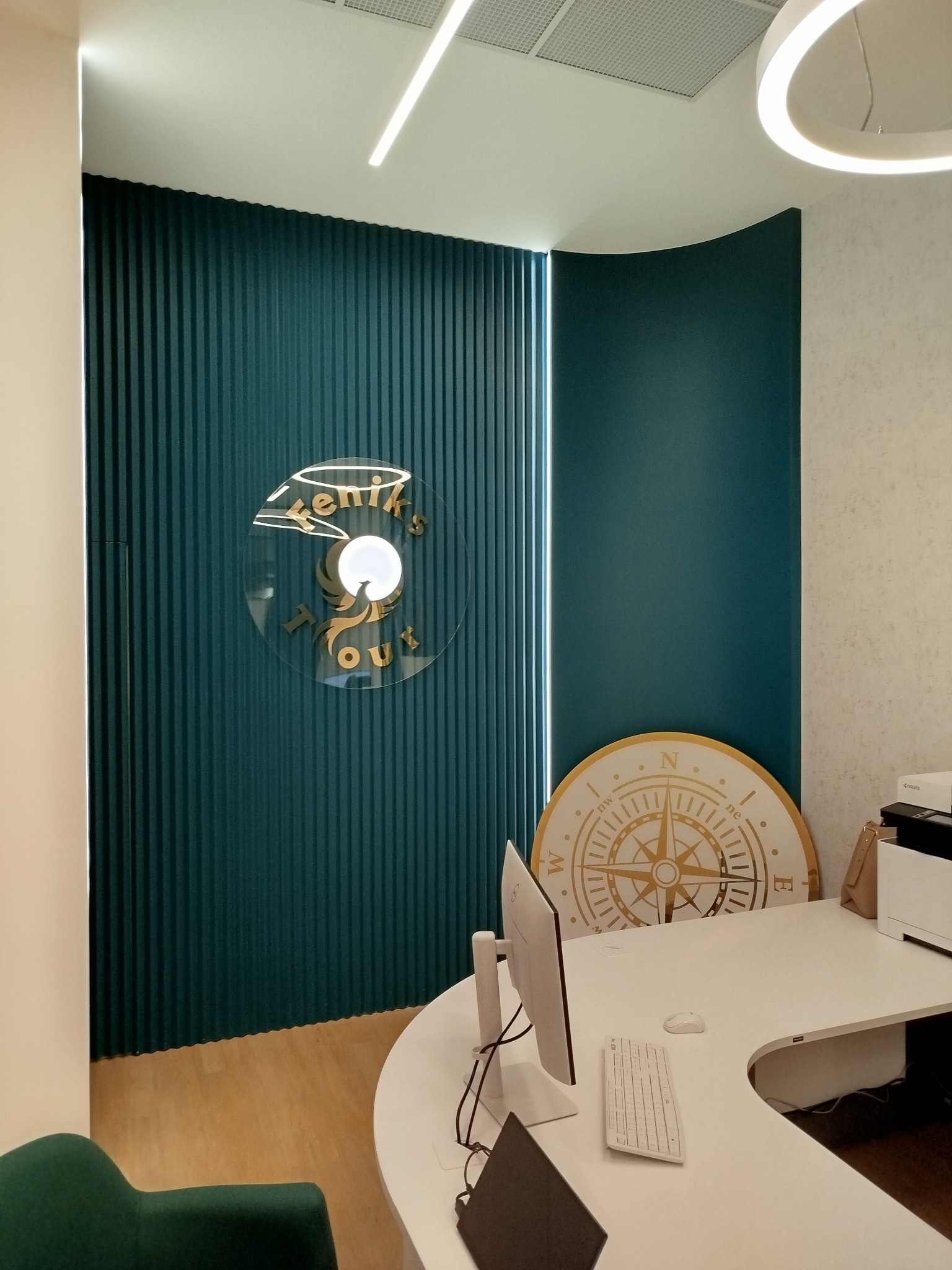 Nowoczesne wnętrze recepcji z turkusową ścianą, logo firmy podróżniczej 'Feniks Tours' i dekoracyjną różą wiatrów, białe biurko z komputerem.