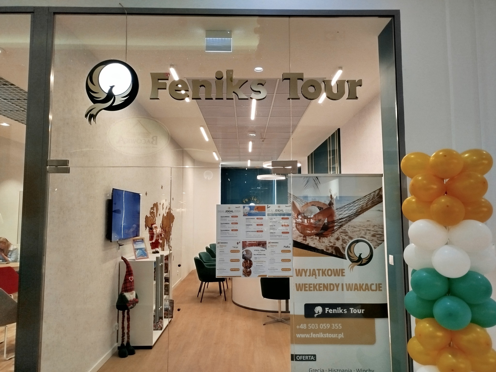 Wejście do biura z logo Feniks Tour, dekoracja z balonów żółto-biało-zielonych, wewnątrz plakaty z ofertami wakacyjnymi i siedzenia dla klientów.