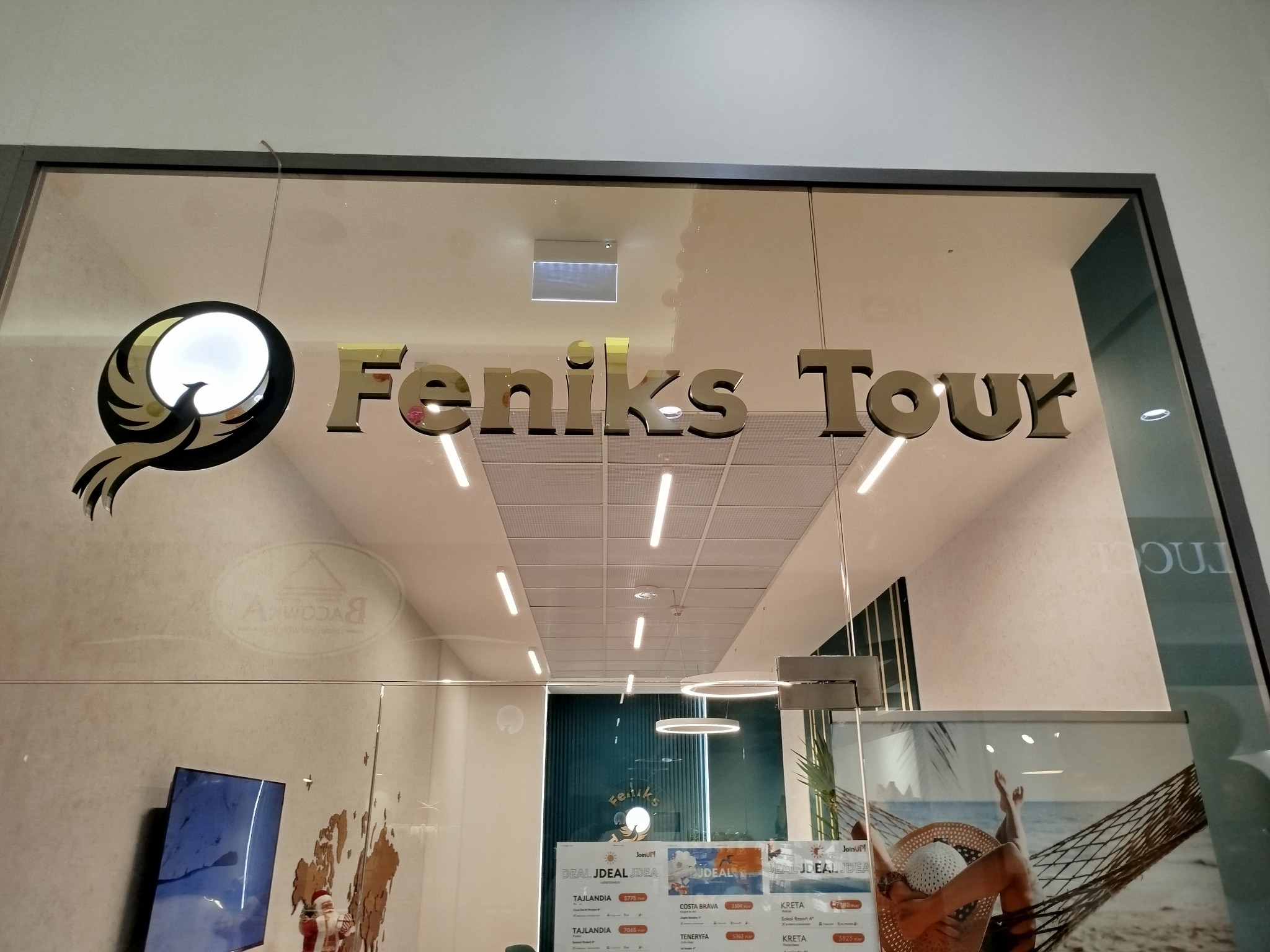 Wejście do biura podróży Feniks Tour z logo złotego feniksa i ofertami wycieczek na plakatach w witrynie.