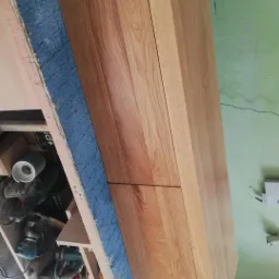 Drewniana komoda z dwiema szufladami, wykonana z jasnego drewna, w warsztacie stolarskim, widoczne narzędzia i akcesoria stolarskie na stole roboczym.