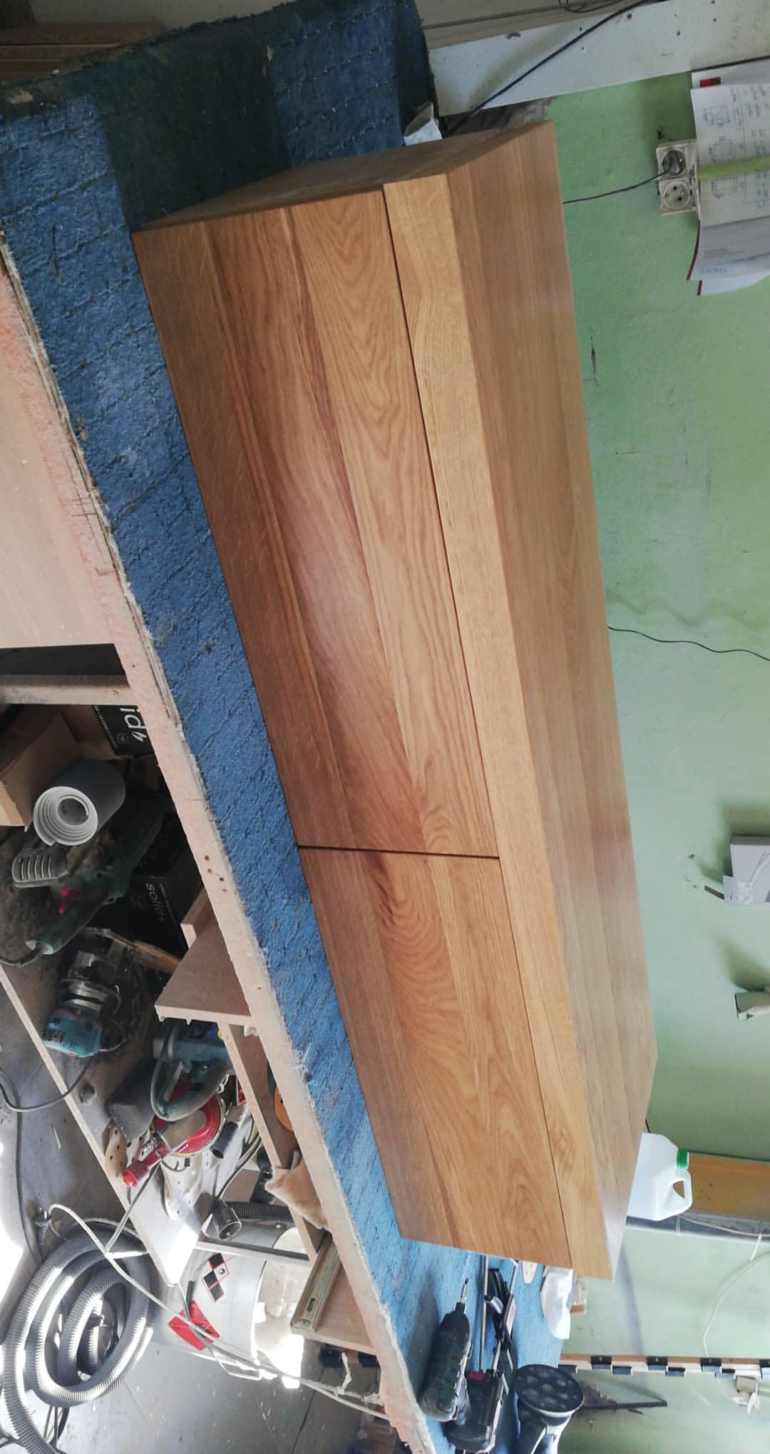 Drewniana komoda z dwiema szufladami, wykonana z jasnego drewna, w warsztacie stolarskim, widoczne narzędzia i akcesoria stolarskie na stole roboczym.