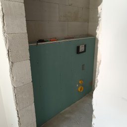 Remonty i wykończenia Ta-bud - Ścianka działowa z zielonej płyty karton gips w trakcie montażu w łazience, widoczne otwory na instalację hydrauliczną i osprzęt, bloczki betonowe i narzędzia na górnej krawędzi płyty.