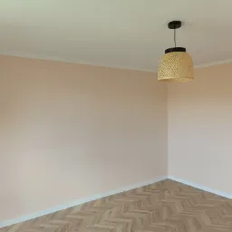 Pomalowane na delikatny róż pokój z parkietem ułożonym we wzór jodełki i wiszącą lampą z wikliny.