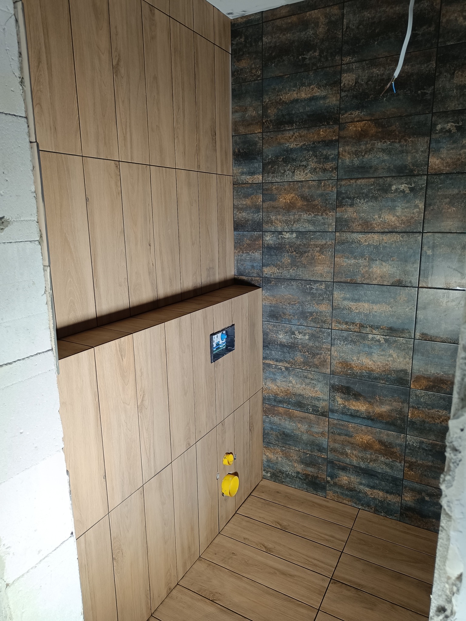 Wykończenie łazienki z drewnopodobnymi płytkami na ścianach i podłodze, widoczne instalacje hydrauliczne i fragment surowej ściany z betonu komórkowego.
