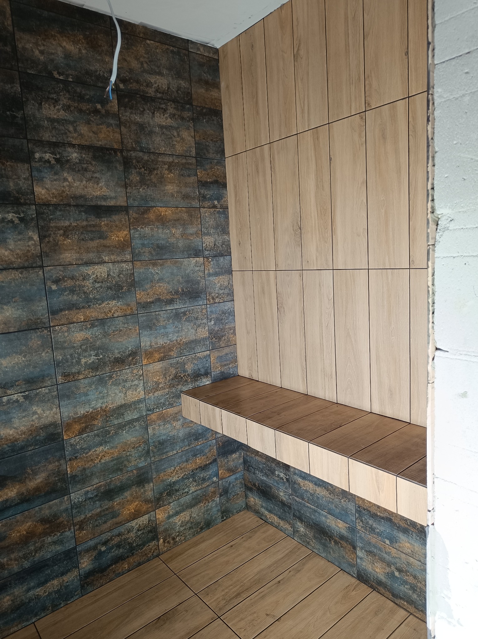 Niedokończona łazienka z drewnopodobnymi płytkami w dwóch odcieniach, z półką z tego samego materiału. Widoczne wystające kable elektryczne i surowy beton.