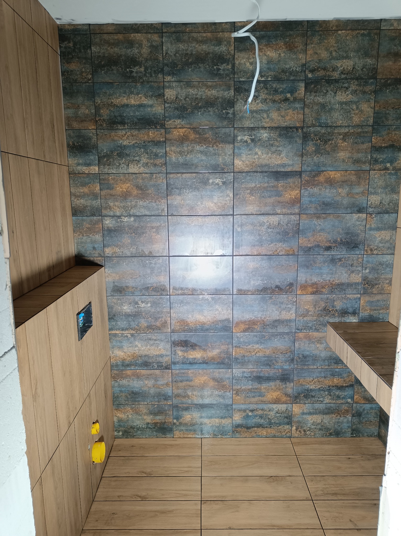 Wykończona łazienka z płytkami imitującymi drewno na podłodze i ścianach, oraz płytkami w odcieniach niebieskiego i brązu na ścianie, widoczne przyłącza hydrauliczne i wystający kabel elektryczny.