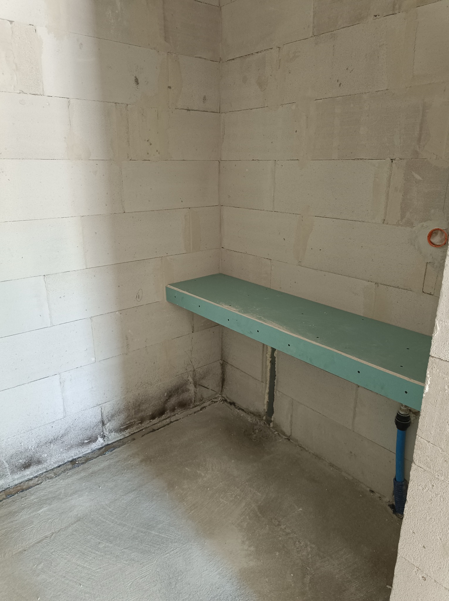 Surowy kąt pomieszczenia z bloczków, z fragmentem zielonej płyty przymocowanej do ściany, widoczne instalacje wodne i ślady wilgoci przy podłodze.