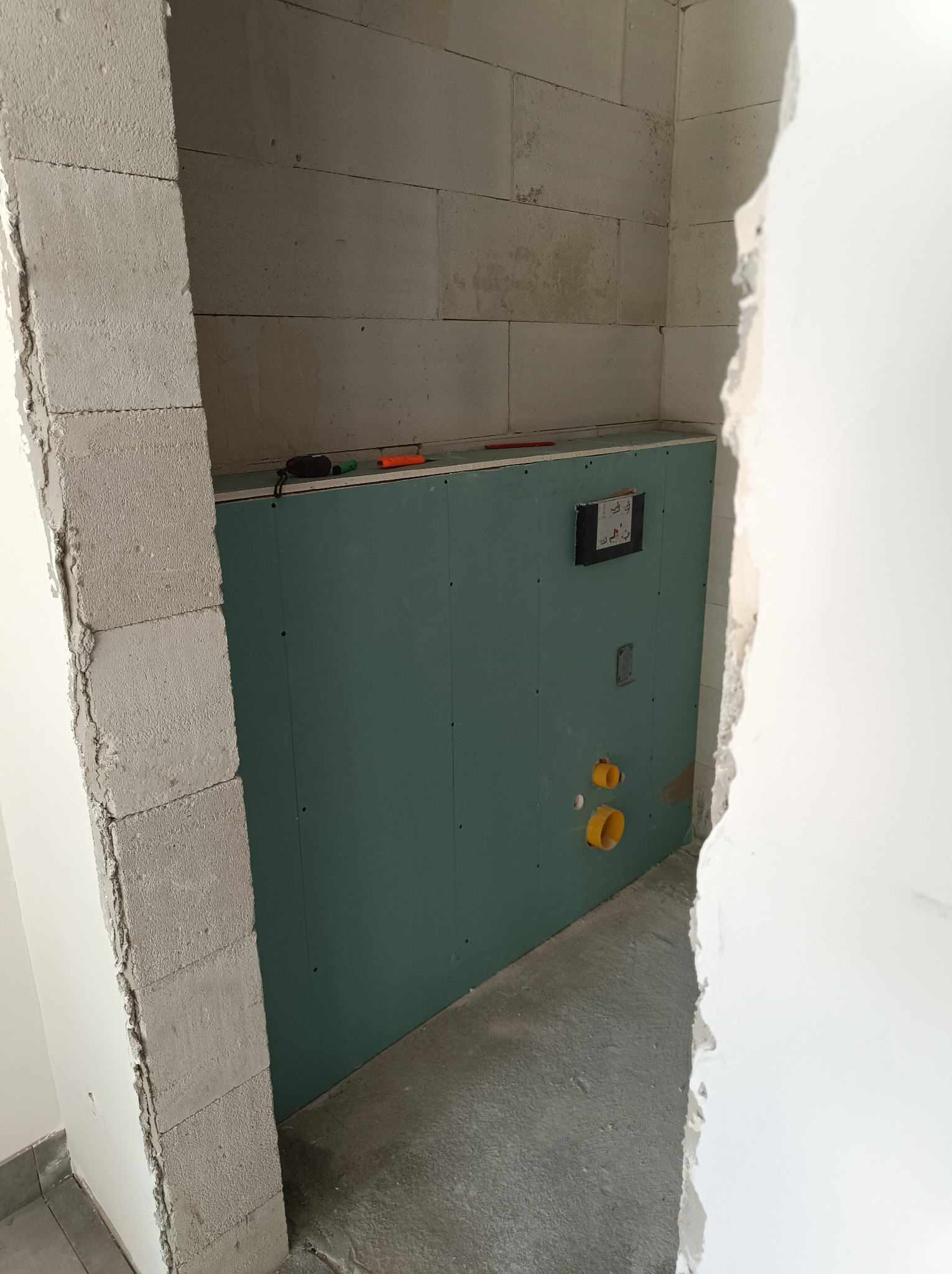 Ścianka działowa z zielonej płyty karton gips w trakcie montażu w łazience, widoczne otwory na instalację hydrauliczną i osprzęt, bloczki betonowe i narzędzia na górnej krawędzi płyty.