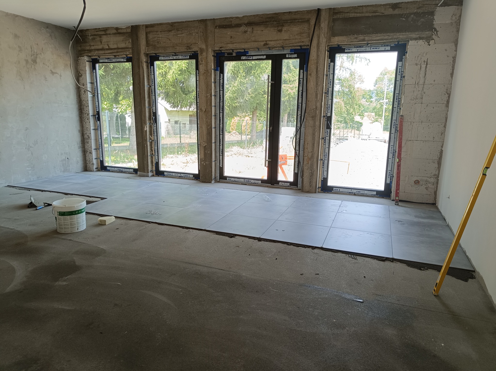 Układanie szarych płytek podłogowych w surowym wnętrzu z trzema oknami balkonowymi, widoczny poziomica i wiadro z klejem.