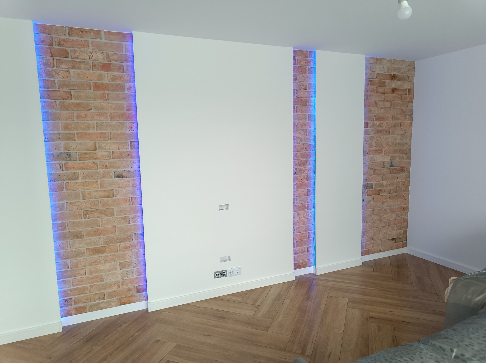 Nowoczesne wykończenie wnętrza z fragmentami ściany z czerwonej cegły, podświetlonymi niebieskim światłem LED, w połączeniu z gładką, białą ścianą i parkietem ułożonym w jodełkę.