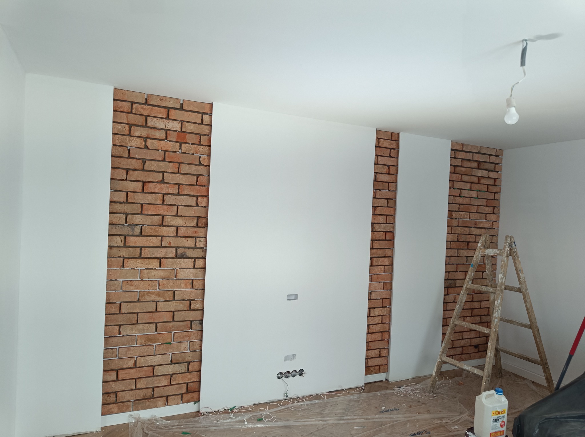 Ściana z fragmentami cegły dekoracyjnej i pomalowanymi na biało panelami, widoczna drabina malarska i butelka z gruntem, podłoga zabezpieczona folią.