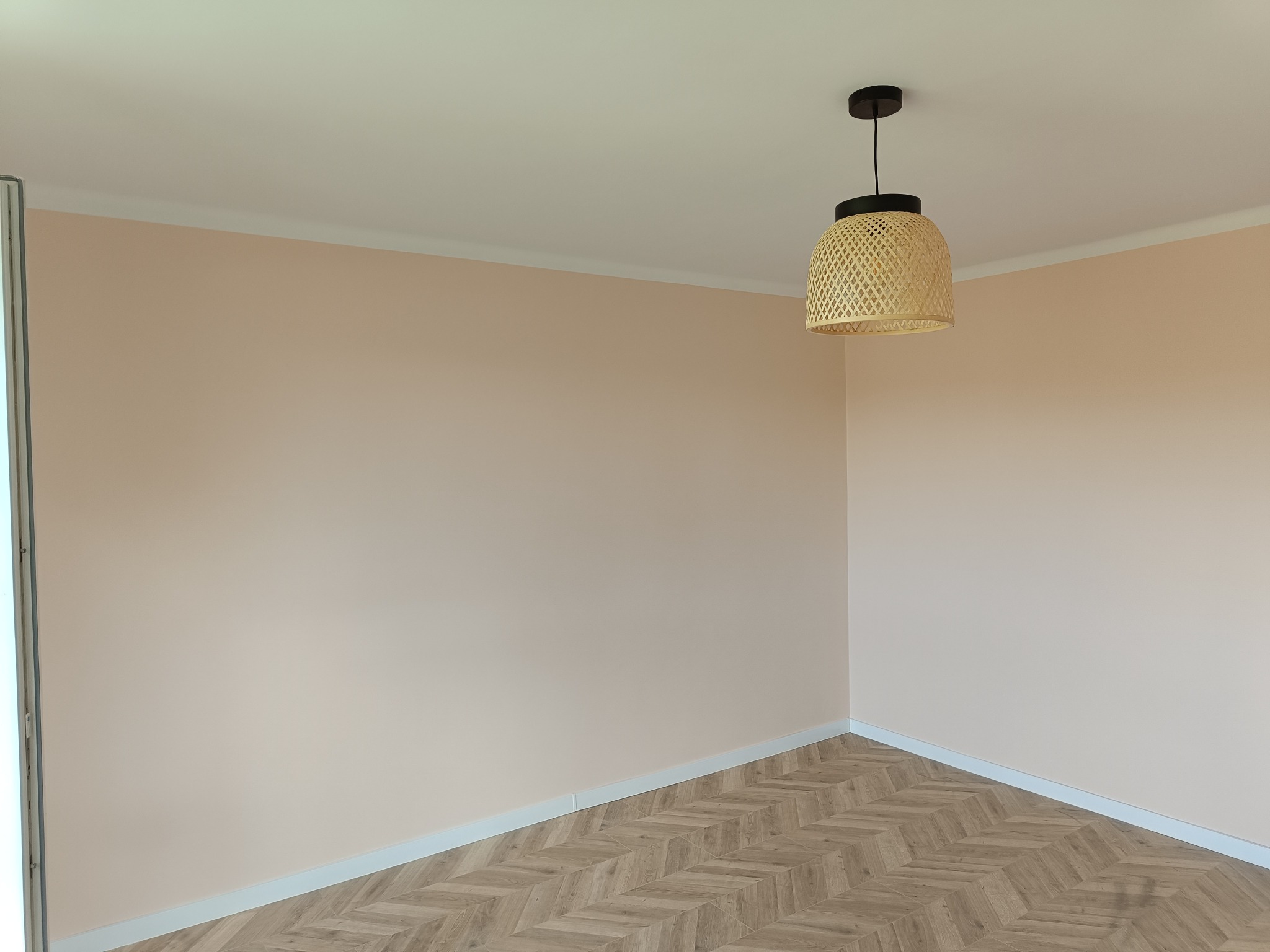 Pomalowane na delikatny róż pokój z parkietem ułożonym we wzór jodełki i wiszącą lampą z wikliny.