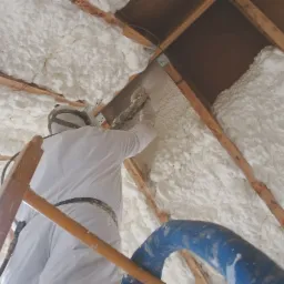 Pracownik w kombinezonie ochronnym aplikuje piankę izolacyjną na poddaszu, widoczne drewniane krokwie i drabina.