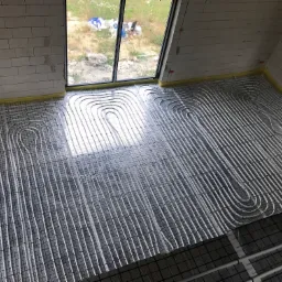 Widok z góry na rozłożone rury ogrzewania podłogowego na siatce montażowej w surowym wnętrzu z oknami i widokiem na zewnątrz.