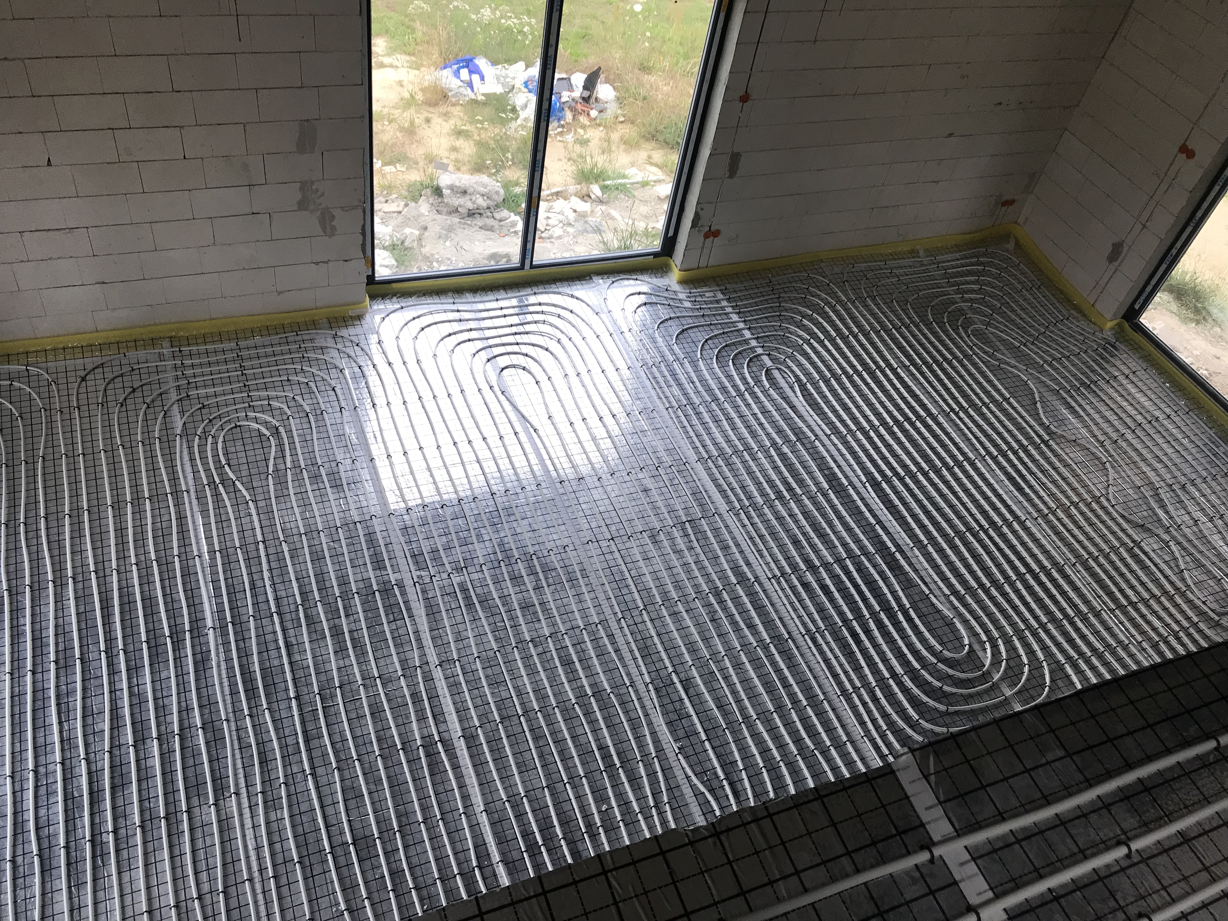 Widok z góry na rozłożone rury ogrzewania podłogowego na siatce montażowej w surowym wnętrzu z oknami i widokiem na zewnątrz.