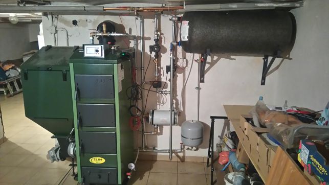 Zielony piec z podajnikiem na ekogroszek, instalacja z rur miedzianych, zbiornik akumulacyjny i osprzęt w kotłowni z widocznymi śladami użytkowania.