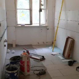 Wnętrze łazienki w trakcie układania białych płytek ceramicznych na ścianach i podłodze; widoczne narzędzia, poziomica, wiadro z zaprawą i paczka kleju ARDEX X6.