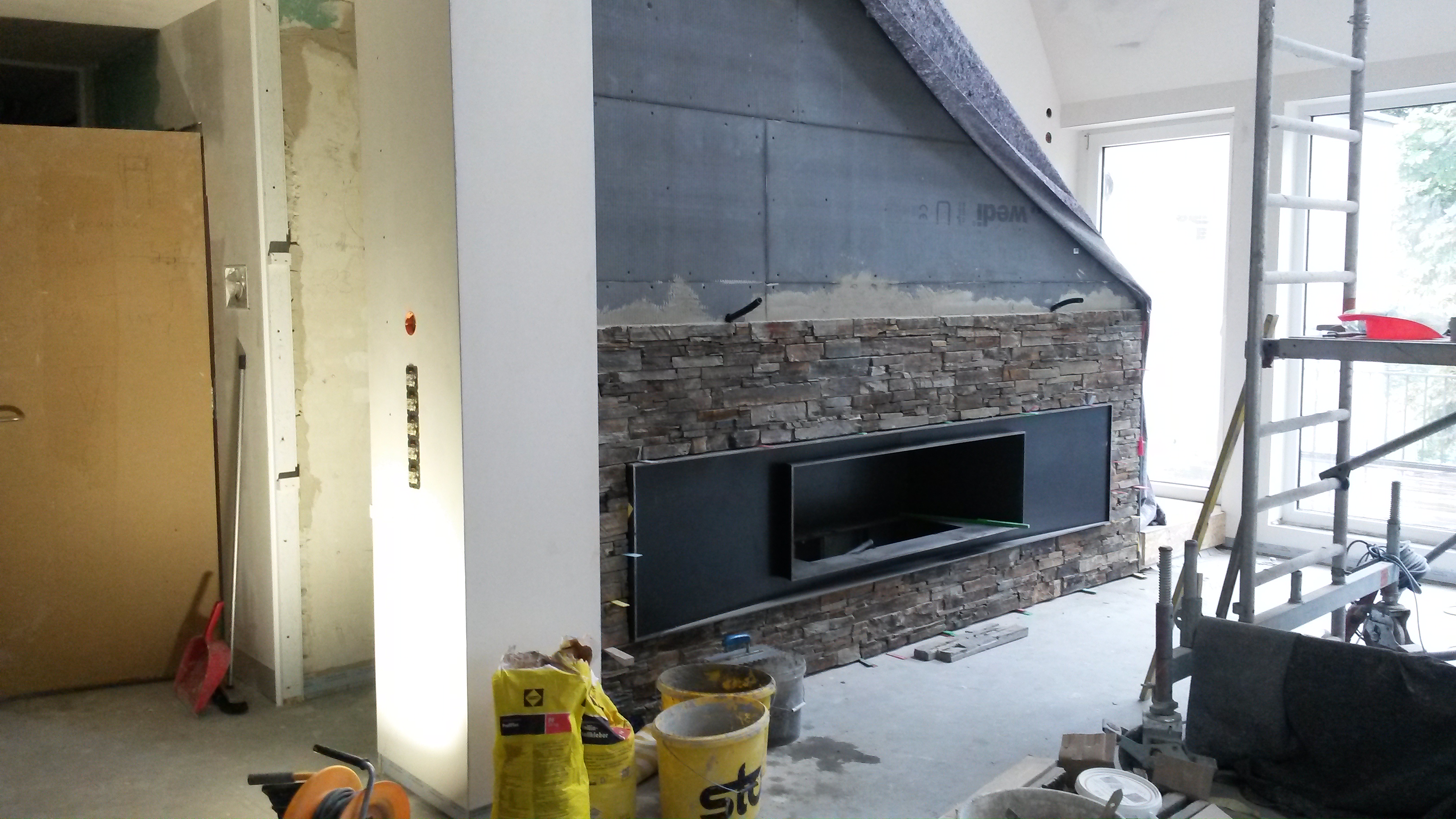 Montaż kamienia dekoracyjnego wokół wnęki na kominek, widoczne płyty izolacyjne Wedi na górze, worki z klejem i drabina w tle.