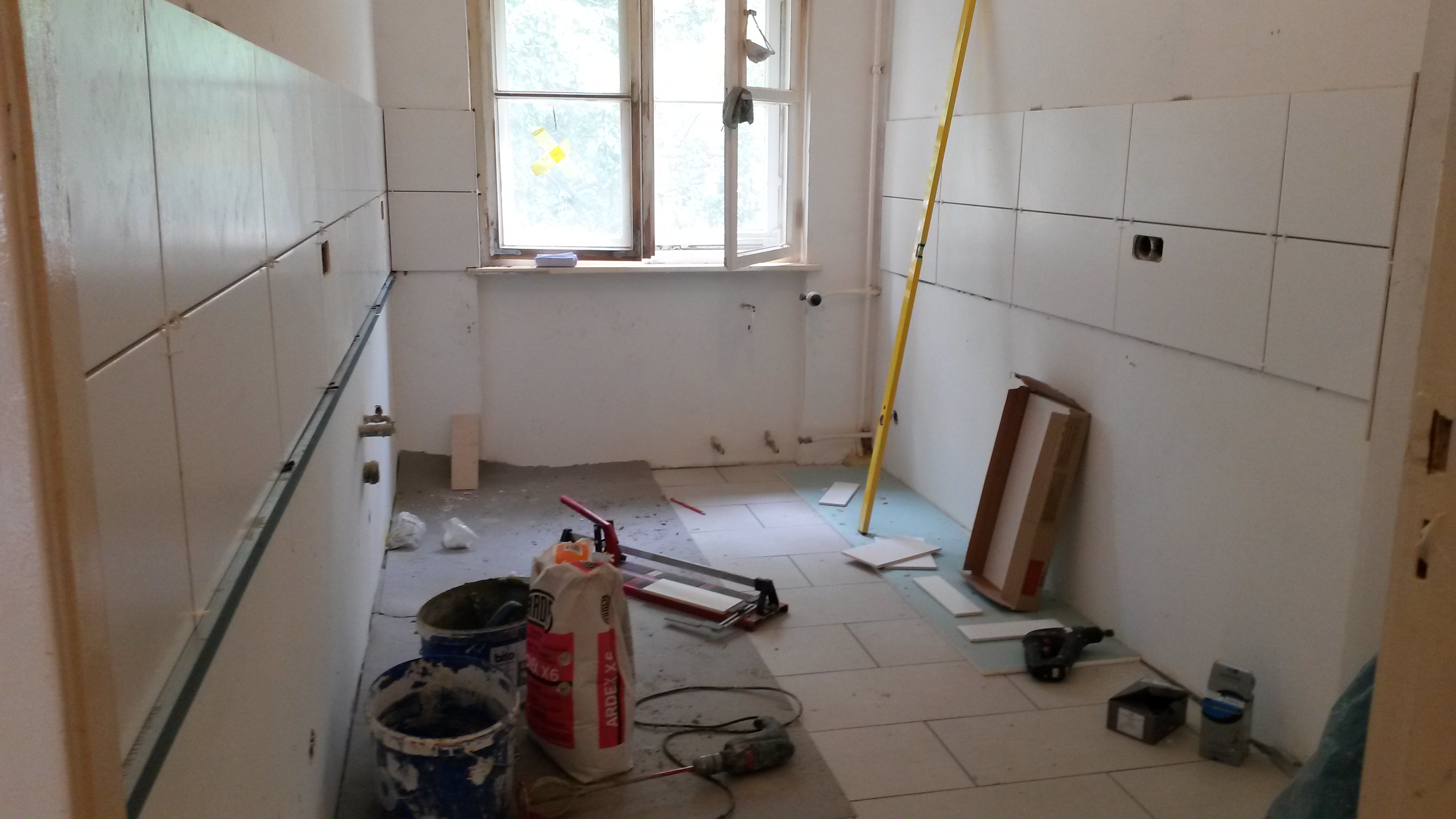 Wnętrze łazienki w trakcie układania białych płytek ceramicznych na ścianach i podłodze; widoczne narzędzia, poziomica, wiadro z zaprawą i paczka kleju ARDEX X6.