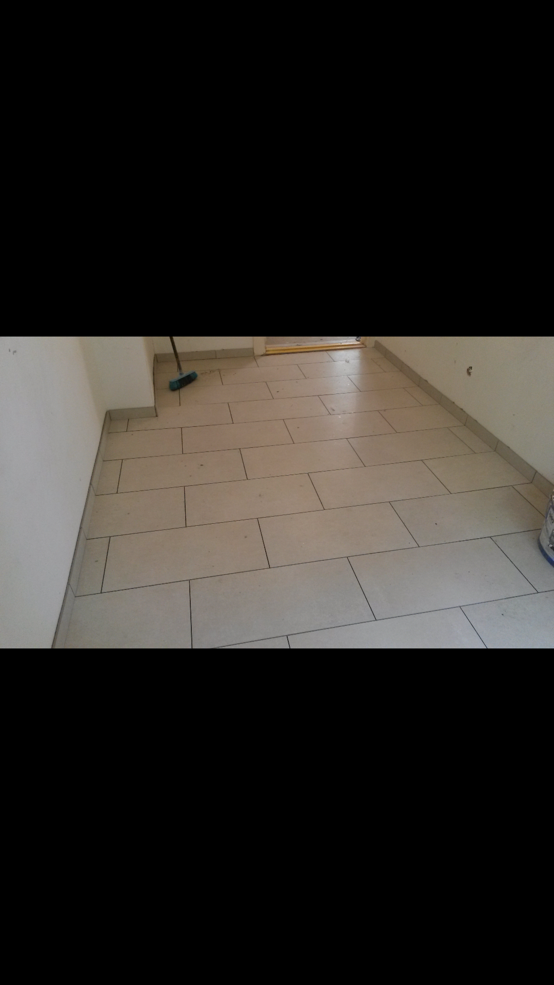 Ułożona podłoga z jasnych, prostokątnych płytek ceramicznych z ciemną fugą, widoczna szczotka i wiadro w rogu pomieszczenia.