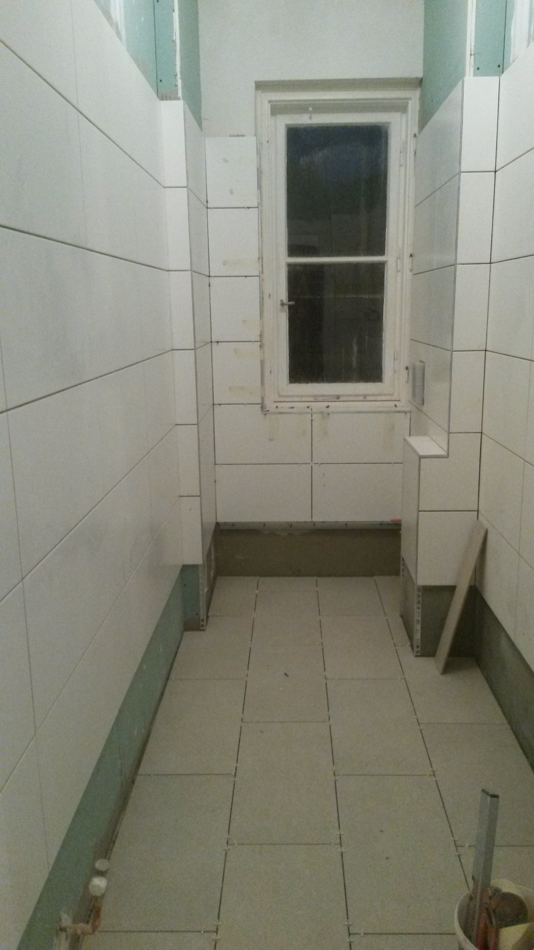 Łazienka w trakcie układania płytek, widoczne białe płytki na ścianach i szare na podłodze, okno w głębi pomieszczenia, brak fug, widoczne narzędzia i materiały budowlane.