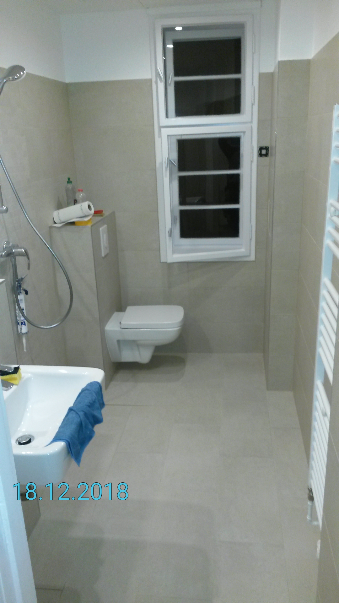 Wykończona łazienka w odcieniach beżu z podwieszaną toaletą, grzejnikiem drabinkowym i oknem z białymi ramami, widok z perspektywy umywalki z niebieskim ręcznikiem.