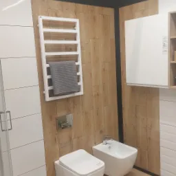 Nowoczesna łazienka z podwieszaną toaletą i bidetem, drewnopodobne płytki na ścianach, biały grzejnik drabinkowy z szarym ręcznikiem, kabina prysznicowa z lewej strony.