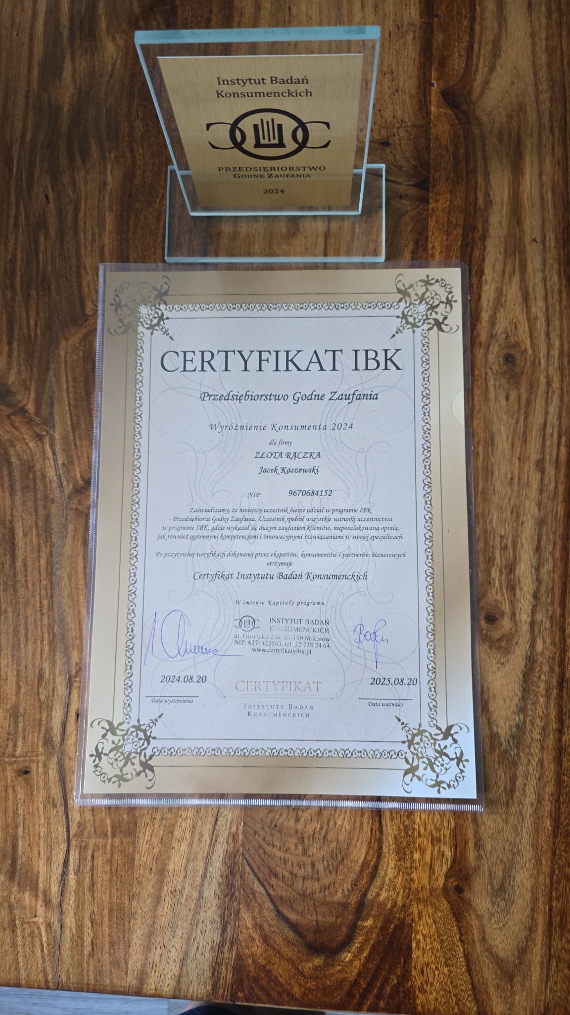 Certyfikat IBK Przedsiębiorstwo Godne Zaufania dla firmy Złota Rączka, umieszczony na drewnianym stole. Nad certyfikatem stoi statuetka Instytutu Badań Konsumenckich.