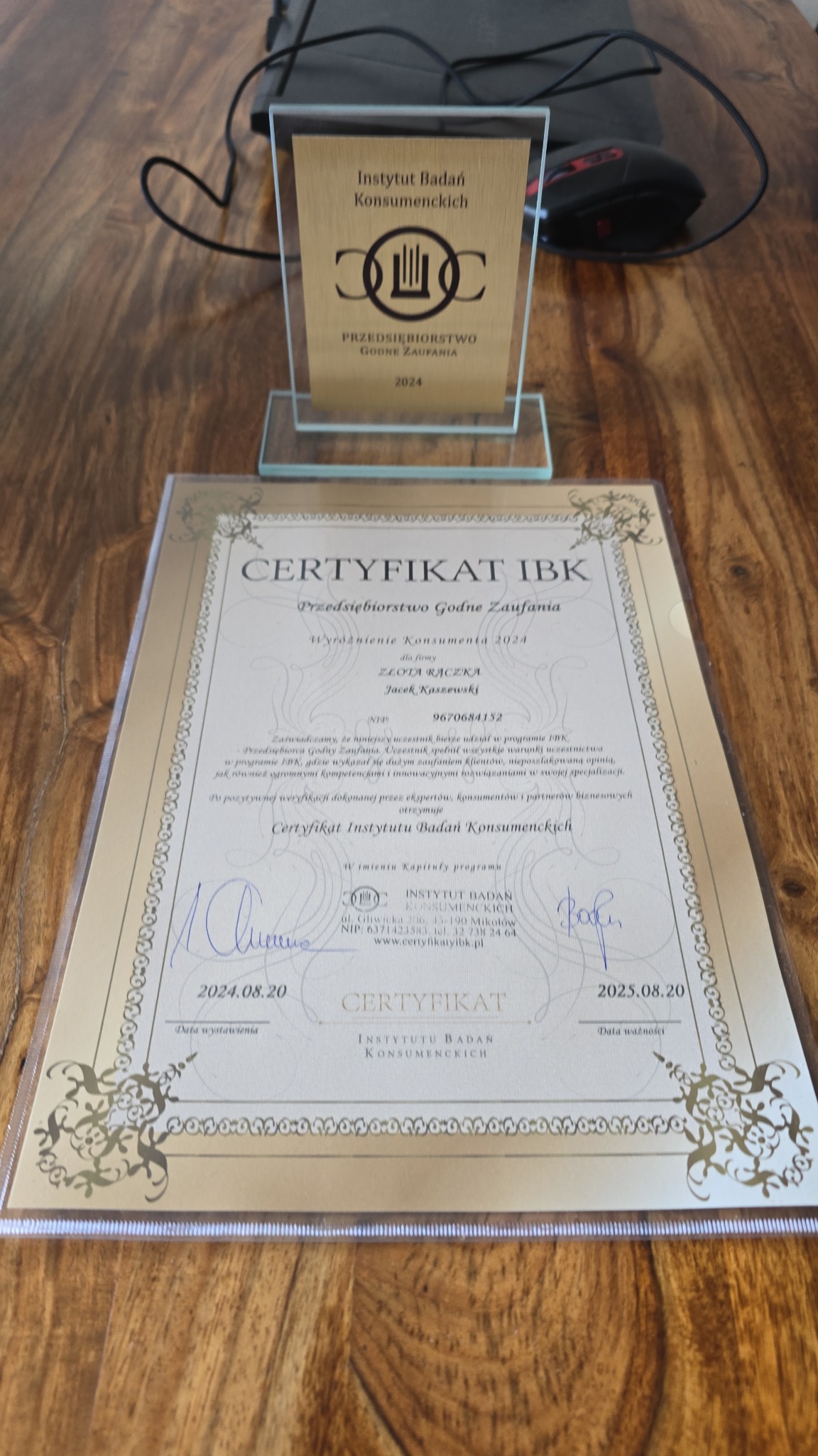 Nagroda i certyfikat 'Przedsiębiorstwo Godne Zaufania 2024' Instytutu Badań Konsumenckich dla firmy 'Złota Rączka' Jacek Kaszewski, leżące na drewnianym stole obok myszki komputerowej.
