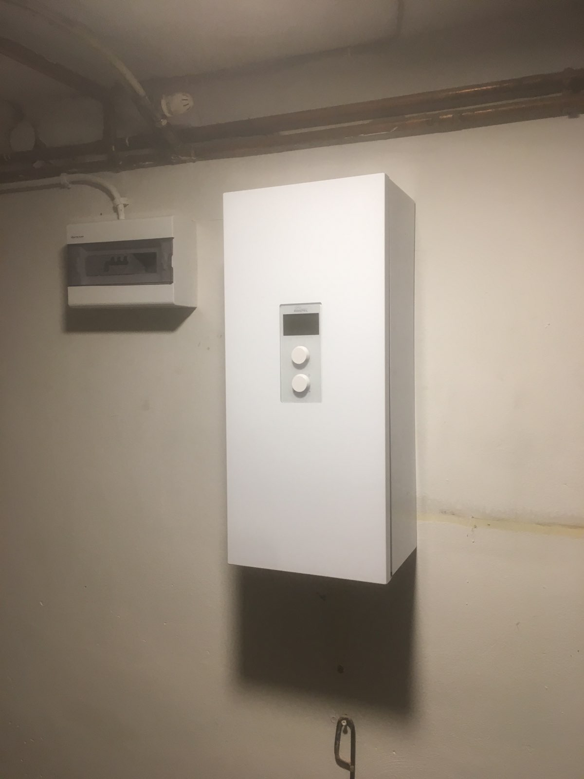 Biała szafka elektryczna z panelem sterowania i skrzynka bezpiecznikowa na ścianie w piwnicy, widoczne rury pod sufitem.