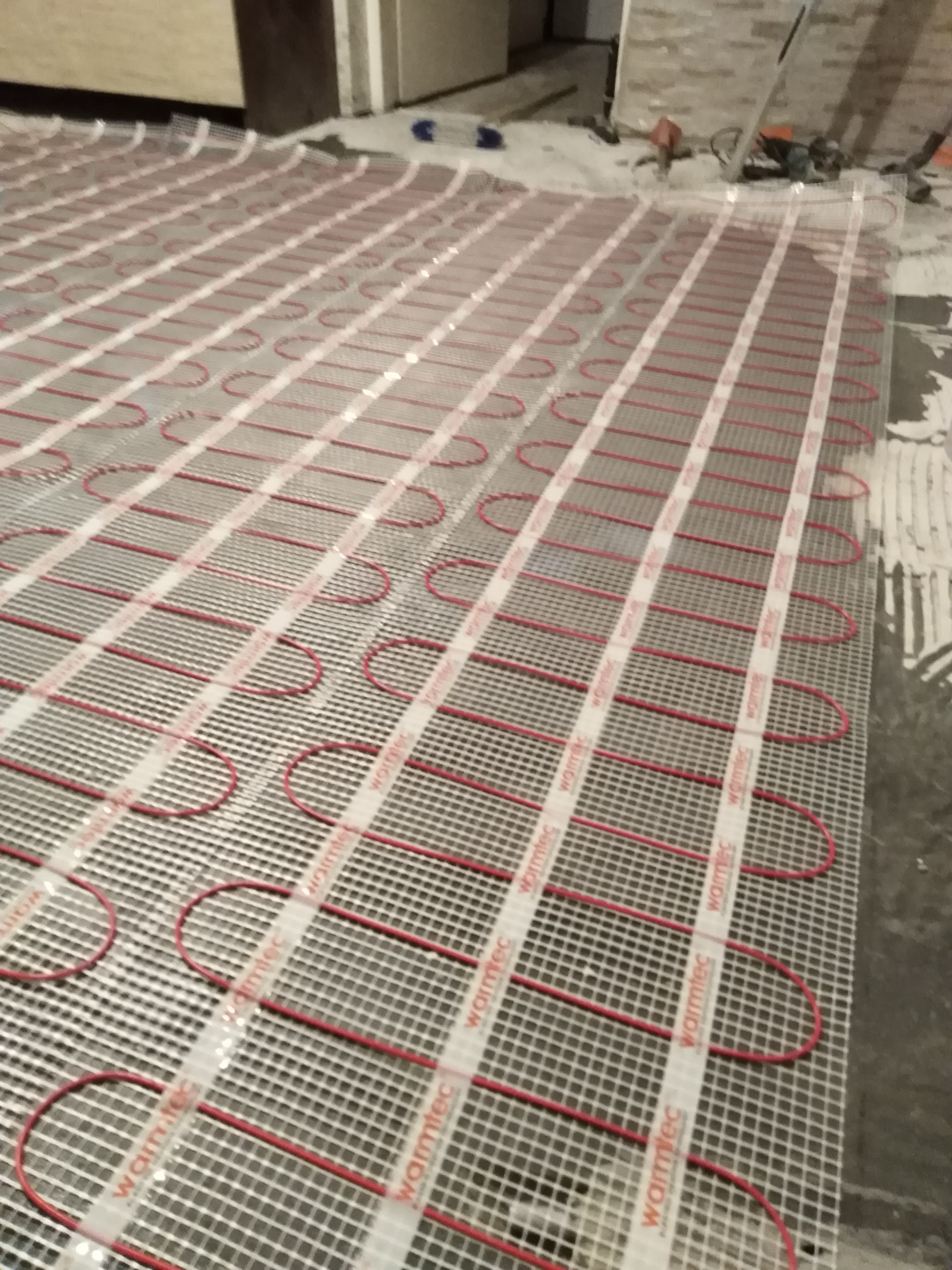 Rozłożona mata grzewcza Warmtec z czerwonymi kablami na podłożu przygotowanym pod płytki, widoczne narzędzia glazurnicze w tle.