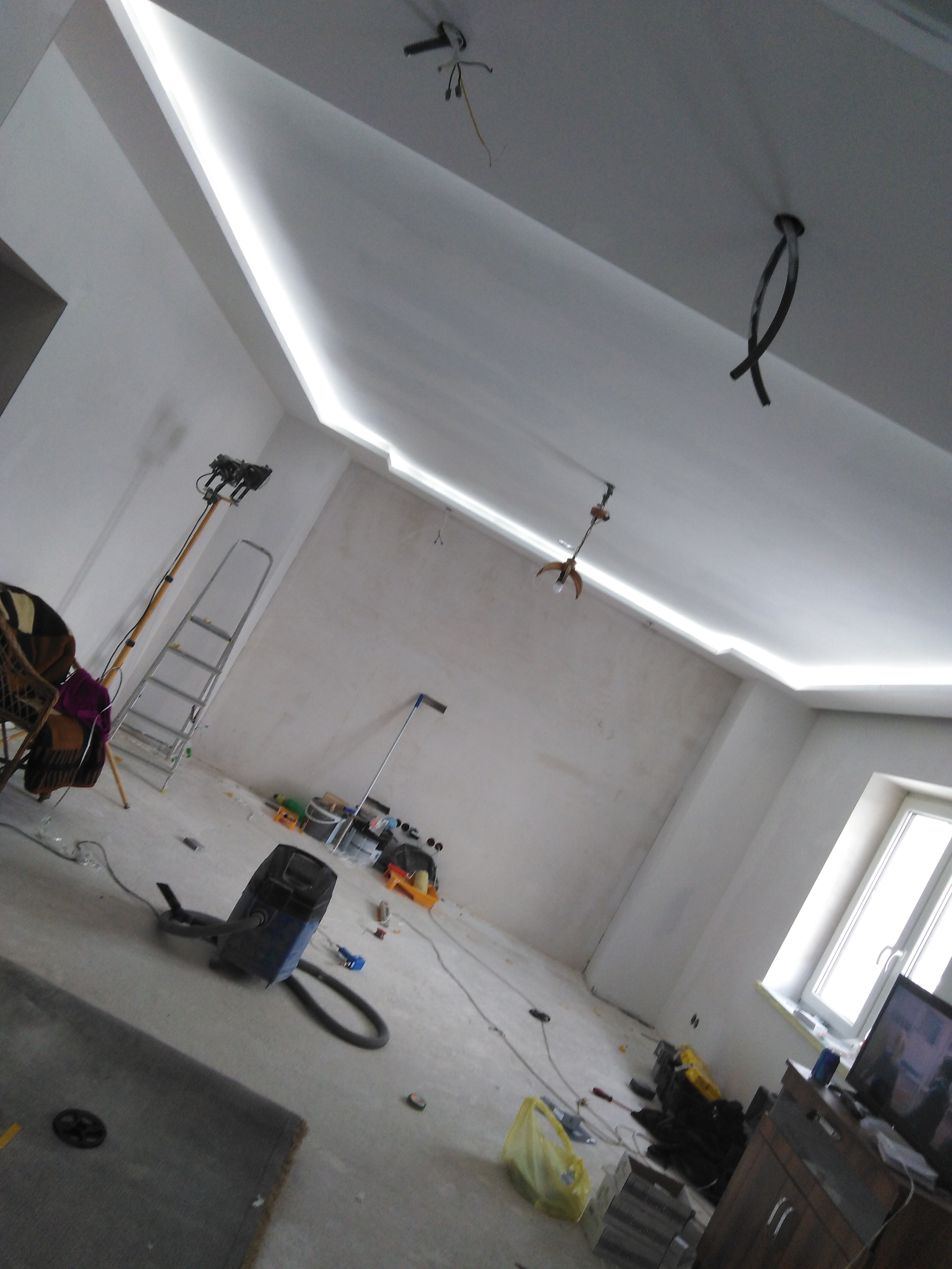 Pokój w trakcie remontu: widoczna surowa ściana, podwieszany sufit z oświetleniem LED, drabina, odkurzacz przemysłowy i narzędzia.