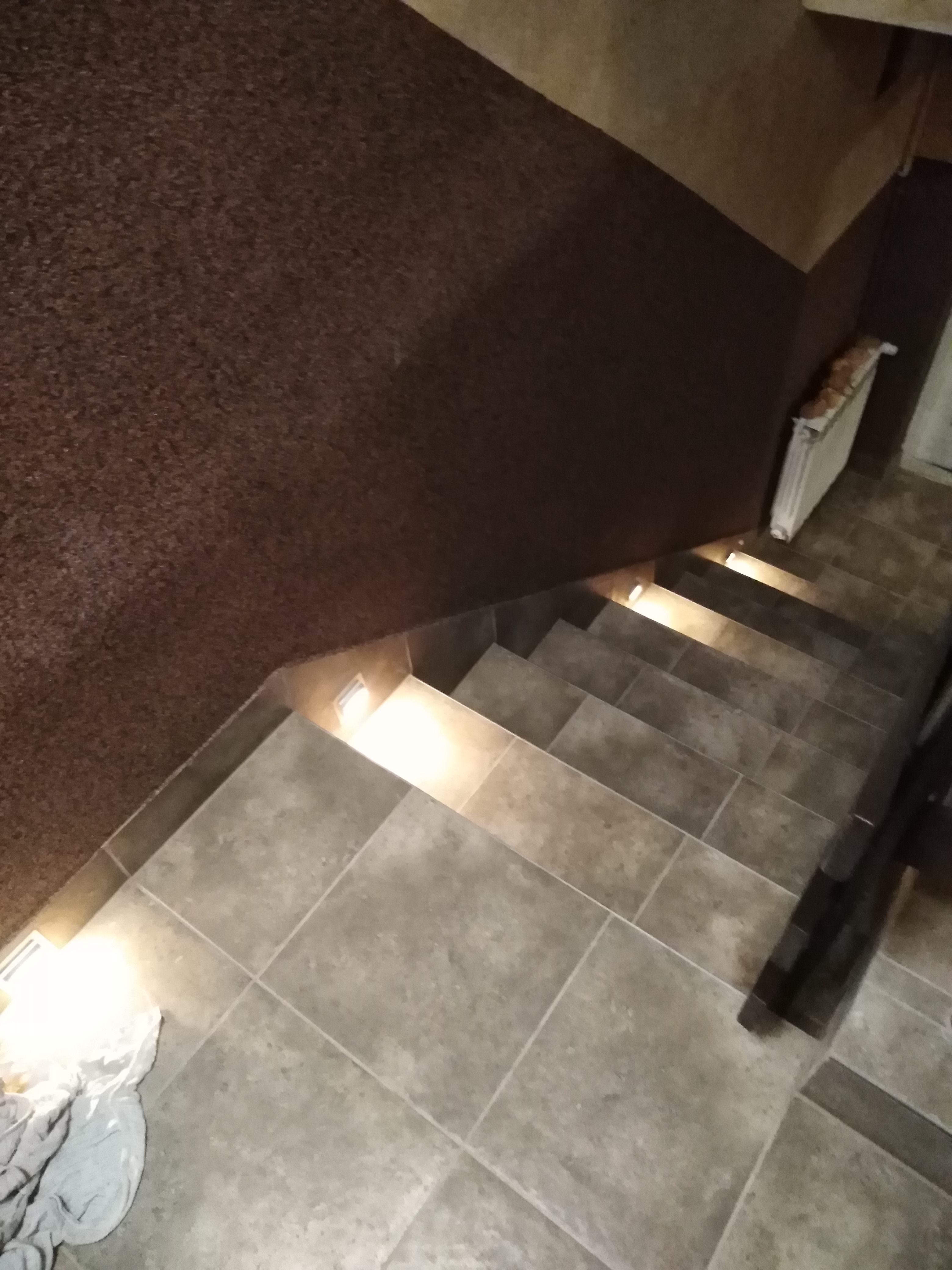 Wykończone schody z szarych płytek, oświetlone małymi, wbudowanymi lampkami. Ciemnobrązowa ściana z fakturą. Widoczny fragment grzejnika. Na dole schodów leży szmatka.
