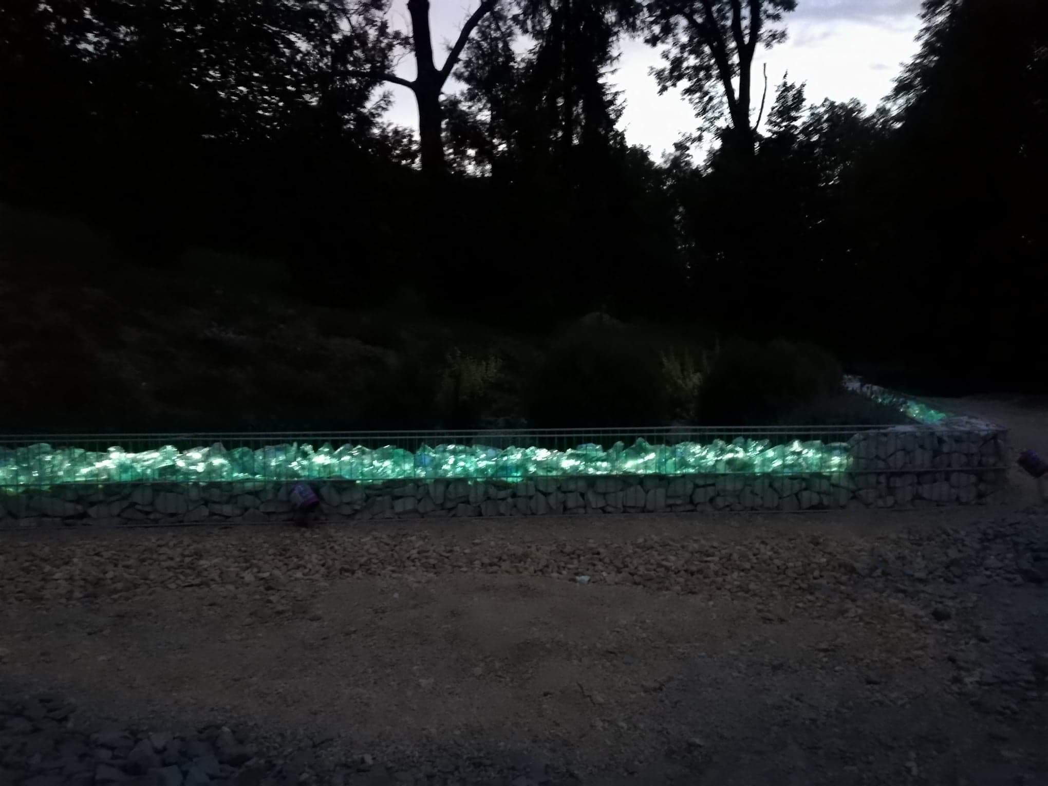 Nocne ujęcie gabionu wypełnionego podświetlonymi, szklanymi bryłami, tworzące efektowną barierę w ogrodzie.